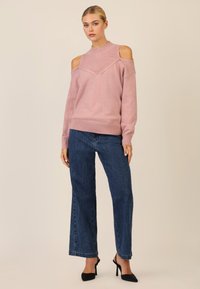Helles rosa gestricktes Sweatshirt mit kalten Schultern, geripptem Kragen und lockerer Passform, kombiniert mit dunkelblauen weiten Jeans und schwarzen High Heels.