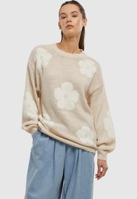 Beige gestrickter Pullover mit großen weißen Blumenmustern. Der lockere Schnitt hat weite Ärmel sowie einen gerippten Kragen und Bündchen.