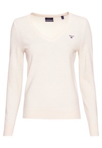 Pull crème en V fabriqué à partir d'un tissu en maille fine, avec des manches longues et un petit logo sur la poitrine gauche. Texture lisse avec des poignets côtelés.
