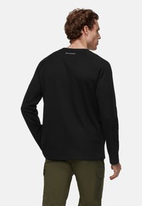 Man wearing black long-sleeve pullover with “MAMMUT” label on upper back, paired with olive green cargo pants, viewed from behind.