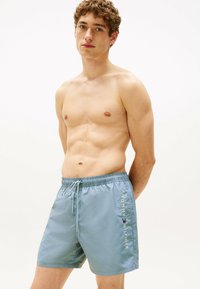 Helle blaue Badeshorts mit elastischem Bund, Kordelzug und Seitentaschen. Das "TOMMY HILFIGER"-Logo in Weiß auf der Seite bedruckt.