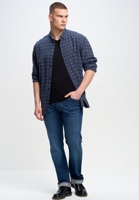 Camisa de cuadros abotonada en azul marino y gris, sobre una camiseta negra. Combinada con jeans azules y zapatos negros, con los puños arremangados.