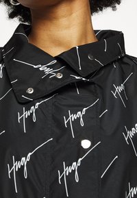 Zbliżenie na kołnierz czarnej kurtki z białym wzorem "Hugo" i srebrnymi zatrzaskami, noszonej przez osobę z ciemnymi kręconymi włosami.