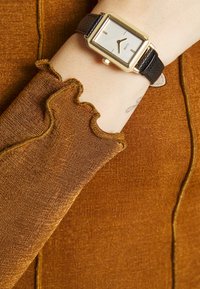 Goldfarbener rechteckiger Uhr mit silbernem Zifferblatt an einem schwarzen strukturierten Lederarmband, kombiniert mit einem rostfarbenen, gerippten Stoffärmel mit gewellten Kanten.