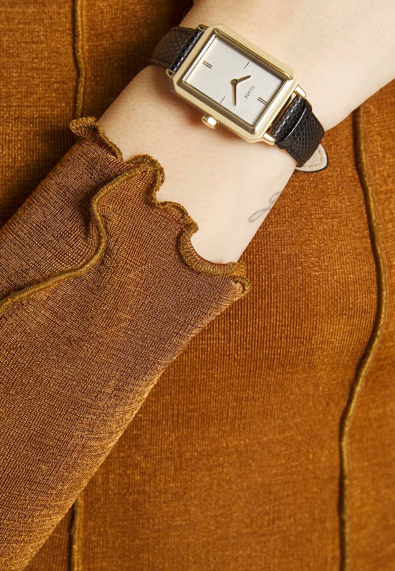 Goldfarbener rechteckiger Uhr mit silbernem Zifferblatt an einem schwarzen strukturierten Lederarmband, kombiniert mit einem rostfarbenen, gerippten Stoffärmel mit gewellten Kanten.