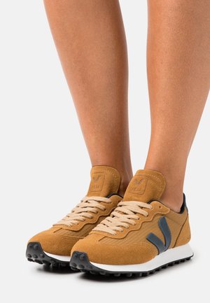 Brązowe zamszowe trampki o fakturowanym wykończeniu, jasnych sznurowadłach, ciemnoniebieskich akcentach i białej podeszwie, z logo na języku.