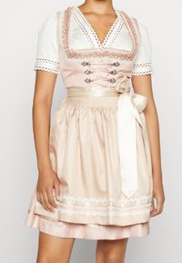 Jasnoróżowa sukienka dirndl z białą bluzką, koronkowym wykończeniem oraz dekoracyjnym sznurowaniem na górze. Satynowa wstążka w talii; kwiatowe koronkowe akcenty na spódnicy.