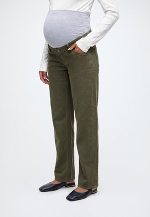 Pessoa grávida a usar calças de maternidade verde-oliva com cintura elástica sobre a barriga, topo branco de manga comprida e sapatos pretos sem fecho.