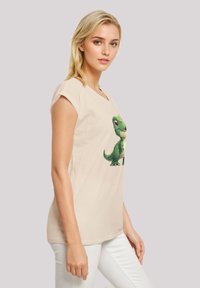 Cream-colored cotton t-shirt with short sleeves featuring a green dinosaur graphic on the front. Smooth texture, relaxed fit.
