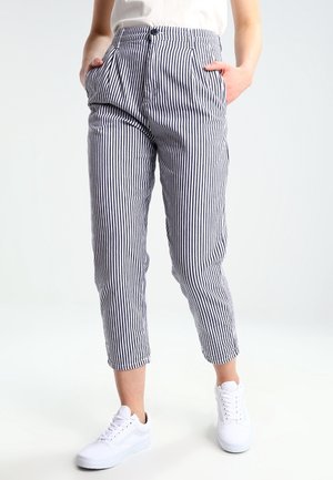 Persona con pantalones a rayas verticales azul marino y blanco, con las manos en los bolsillos y zapatillas blancas, de pie sobre una superficie blanca.