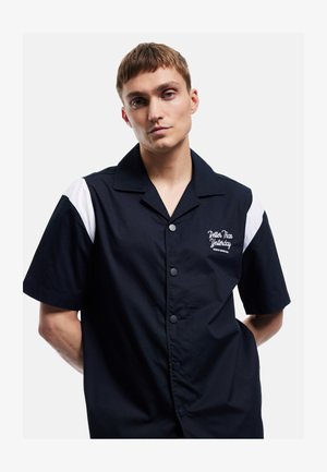 Jonge man draagt een zwart overhemd met korte mouwen, witte accenten op de schouders en de tekst "Better Than Yesterday" op de borst.