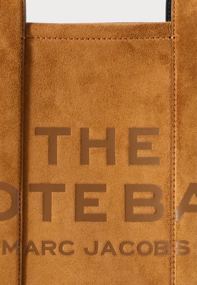 Négyzetes suéd táska, amelyen nagy dombornyomott felirat olvasható: "A TÁSKA" és "MARC JACOBS". Sima felület, letisztult vonalak és minimalista dizájn jellemzi.