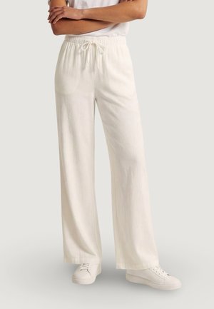 Persona che indossa pantaloni bianchi larghi con coulisse e sneakers bianche, in piedi con le braccia incrociate davanti a uno sfondo semplice.