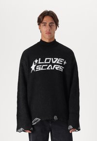 Μαύρος πλεκτός σκούφος με ψηλό γιακά, που διαθέτει λευκό κείμενο "LOVE SCARS" και γραφικά με αστέρια. Με φθαρμένες άκρες και χαλαρή εφαρμογή, υφασμάτινη υφή.