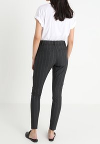 Žena nosící úzké kalhoty s tenkými šedými proužky, bílým tričkem s ohrnutými rukávy a černými nazouvacími botami, stojící zády.