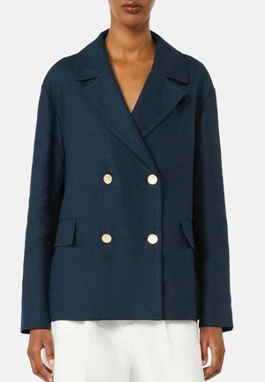 Donna che indossa una giacca doppiopetto blu navy con bottoni dorati e tasche con pattina, abbinata a pantaloni bianchi.