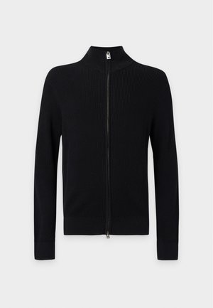 Jeune homme portant un pull côtelé noir à fermeture éclair, un pantalon foncé et des chaussures noires, debout devant un fond clair uni.