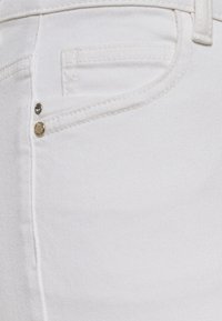 Tissu en denim blanc avec une poche et des garnitures dorées. La surface a une texture lisse avec des détails de couture subtils.
