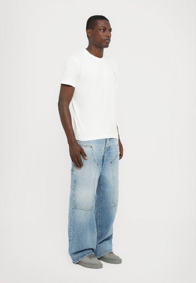 Tricou alb din bumbac asortat cu blugi lejeri de denim de culoare albastră deschisă. Blugii au buzunare cargo și o textură ușor estompată. Adidași gri.