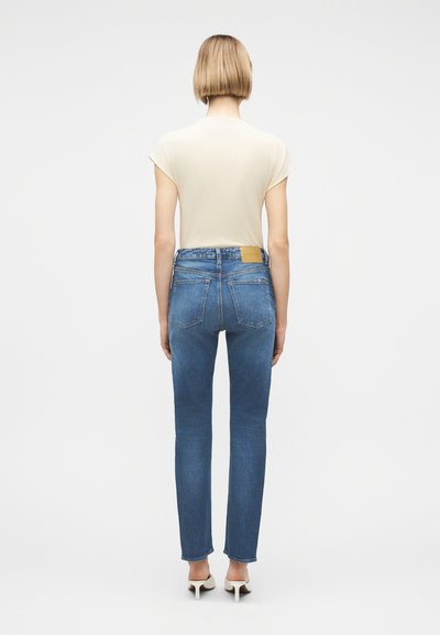 Γυναίκα με κοντά ξανθά μαλλιά στέκεται με την πλάτη προς τα εμπρός, φοράει μπεζ κοντομάνικη μπλούζα, μπλε τζιν ψηλόμεσο και λευκά παπούτσια με τακούνι σε λευκό φόντο.