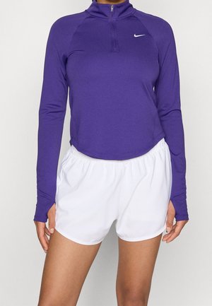 Femme portant un haut de sport à manches longues violet avec une fermeture éclair à l'avant et un short de sport à taille élastique blanche.