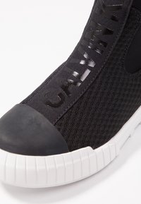 Černá síťová teniska s lesklým označením "CALVIN", gumovým ochranným krytem špičky a bílou texturovanou podrážkou. Vyznačuje se elegantním designem a materiály.