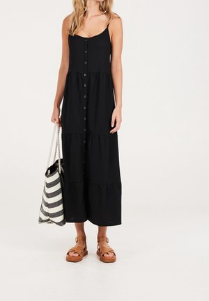 Kobieta w czarnej, bezrękawnej sukience maxi zapinanej na guziki z przodu, brązowych sandałach na platformie oraz trzymająca czarno-białą torbę na ramię w paski.