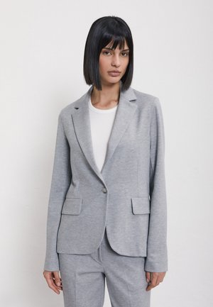 Femme aux cheveux noirs raides en carré, portant une veste de costume gris clair et un pantalon assorti par-dessus un top blanc, posant devant un fond blanc uni.