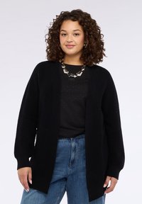 Cardigan nero aperto sul davanti, realizzato in tessuto morbido, con maniche lunghe e vestibilità rilassata. Chi lo indossa lo abbina a una camicia nera e a dei denim.