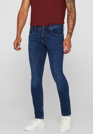 Man draagt slanke donkerblauwe jeans, witte sneakers en een bordeauxrood T-shirt, staand met ontspannen armen langs het lichaam.