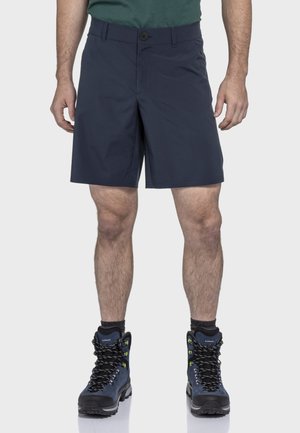 Marineblaue Shorts mit geradem Schnitt, ausgestattet mit einem Knopfverschluss und Taschen. Kombiniert mit schwarzen Wanderschuhen und dunklen Socken.
