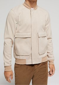 Beige herringbone jacket with two front pockets, ribbed collar and cuffs, and a solid beige waistband. Soft texture, fitted design.