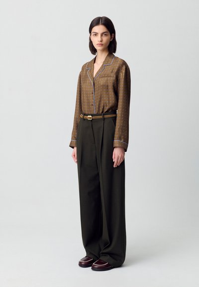 Donna che indossa una camicia a bottone marrone con motivo, infilata in pantaloni a gamba larga con vita alta di colore verde scuro, con una cintura e mocassini scuri.