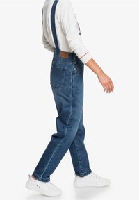 Jeansbyxor i mörkblått med justerbara remmar, knäppning fram och fickor, tillsammans med en vit långärmad topp och vita sneakers.