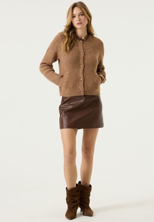 Frau steht in einer braunen Strickjacke, einem dunkelbraunen Minirock aus Leder, schlabbrigen braunen Stiefeln und einer Halskette aus Perlen, die Hände in den Taschen.