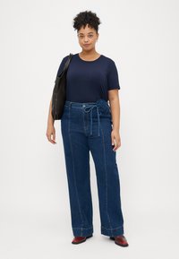 Top de manga curta navy combinado com calças de ganga de cintura alta, com um detalhe em laço floral, complementado por uma bolsa tote preta e sapatos vermelhos.