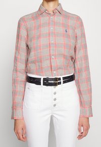 Rutig knyt skjorta i röda och grå nyanser, med klassisk krage och långa ärmar. Bärs med ett svart flätat bälte och vita högmidjade jeans.