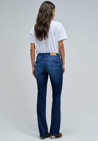 Mujer con cabello largo castaño que lleva una camiseta blanca, jeans bootcut oscuros y zapatos de tacón marrones, de pie frente a un fondo simple.