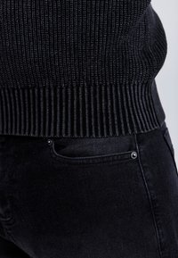 Fekete bordázott pulóver texturált felülettel, sötét farmerrel párosítva, amelyen egy kis ezüst gomb található a zsebnél, és sima anyagú.