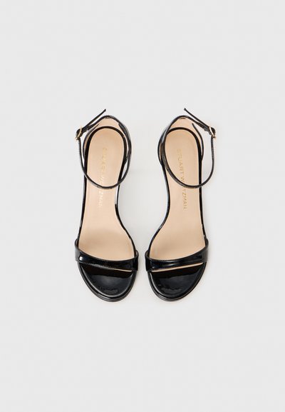 Sandales ouvertes en cuir verni noir avec des brides de cheville et une boucle en or, semelle plate, montrées de dessus sur fond blanc.