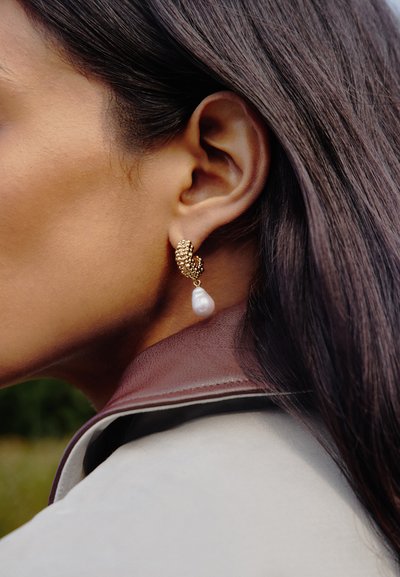Nahaufnahme eines Ohrs einer Person, die einen goldfarbenen, strukturierten Creolen-Ohrring mit einer hängenden weißen Perle trägt, dunkles Haar und ein beiger Jackenkragen sind sichtbar.
