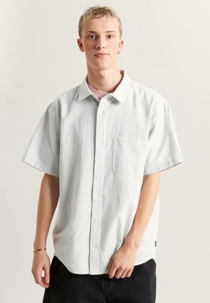 Camisa de botones de manga corta a rayas en azul claro y blanco, con un bolsillo en el pecho y un corte holgado. La tela parece suave y similar al algodón.