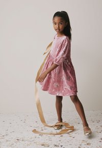 Roze fluwelen jurk met pofmouwen en bloemmotieven, gecombineerd met gouden ballerina's en een lange gouden lint vasthoudend.