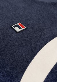 Tissu texturé bleu marine avec un petit patch logo brodé présentant un design en noir, blanc et rouge, ainsi qu'une bande blanche courbée.