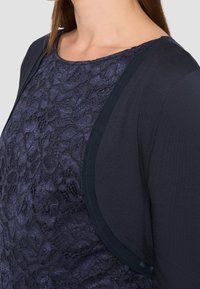 Woman wearing a navy lace top with a sheer floral pattern and a dark navy open cardigan.