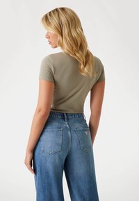 T-shirt verde oliva aderente con maniche corte, abbinato a jeans azzurri a vita alta con tasche applicate e etichetta del marchio sul retro.