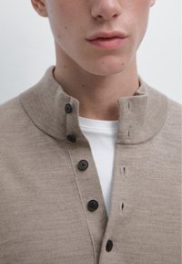 Maglione beige lavorato a maglia con colletto alto e tre bottoni neri. Indossato sopra una t-shirt bianca, presenta una texture morbida e un design semplice.