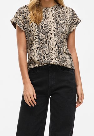 Kvinde iført en kortærmet beige t-shirt med slangeprint, stukket ned i højtaljede sorte bukser med hænderne afslappet ned langs siderne.