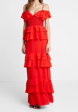 Mujer vestida con un vestido rojo hasta el suelo con volantes en capas, tirantes finos y mangas con volantes fuera del hombro, de pie sobre un fondo blanco.