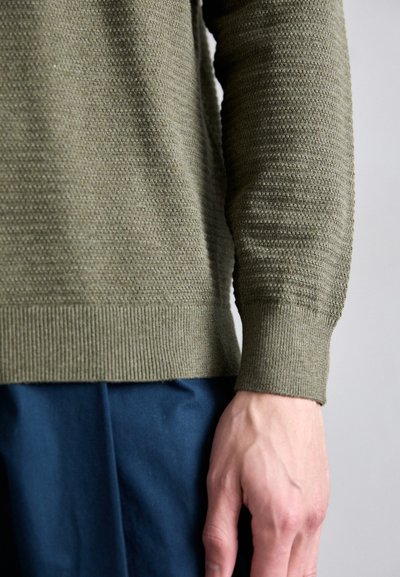 Pull en vert olive texturé avec des poignets côtelés, gros plan sur le tissu mettant en valeur des motifs horizontaux et une finition lisse. Pantalon bleu partiellement visible.
